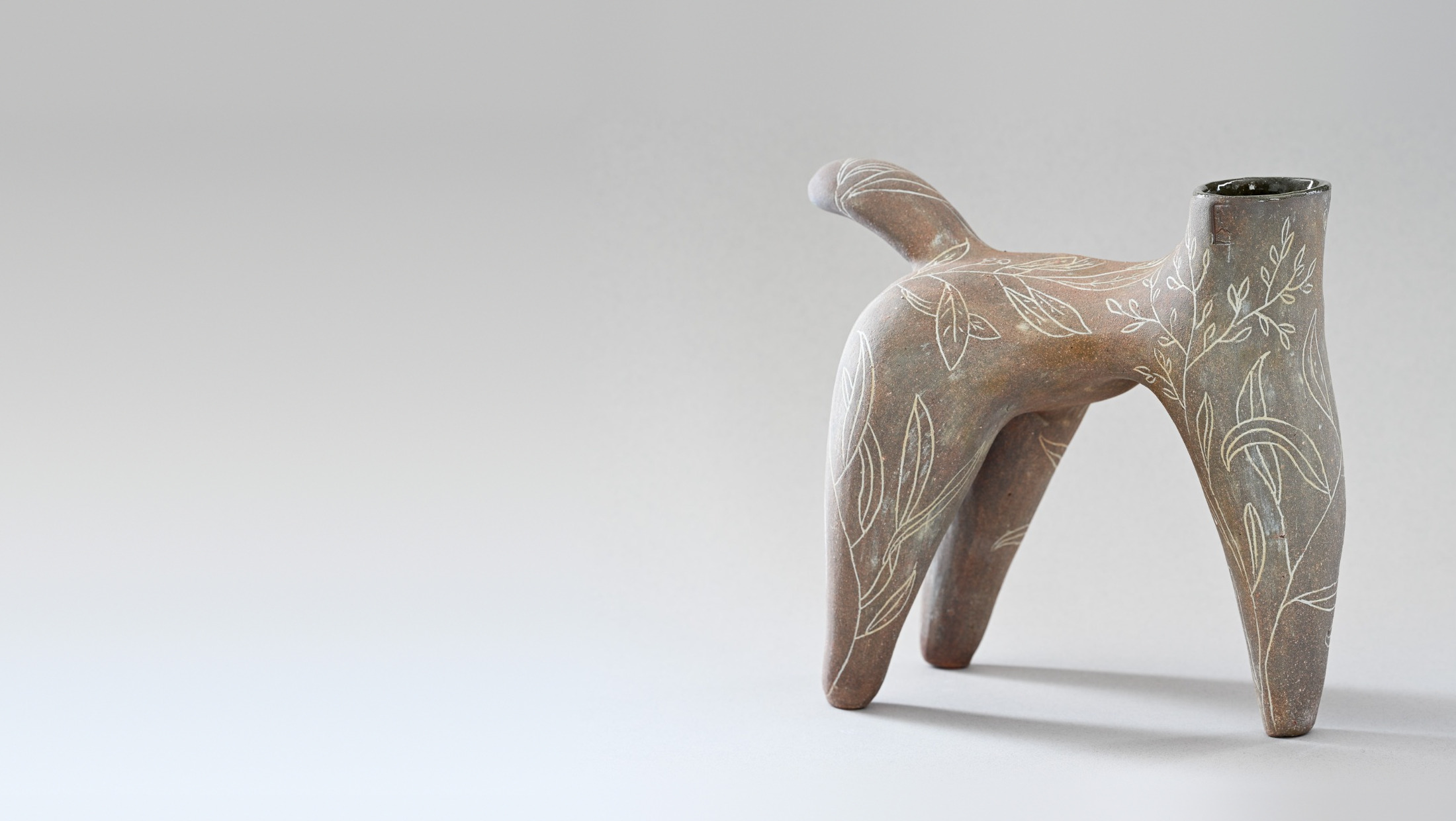 La pièce entière : sculpture en terre à quatre pieds, au long cou ouvert, couverte de feuillages incisés en blanc, sur fond gris de studio.