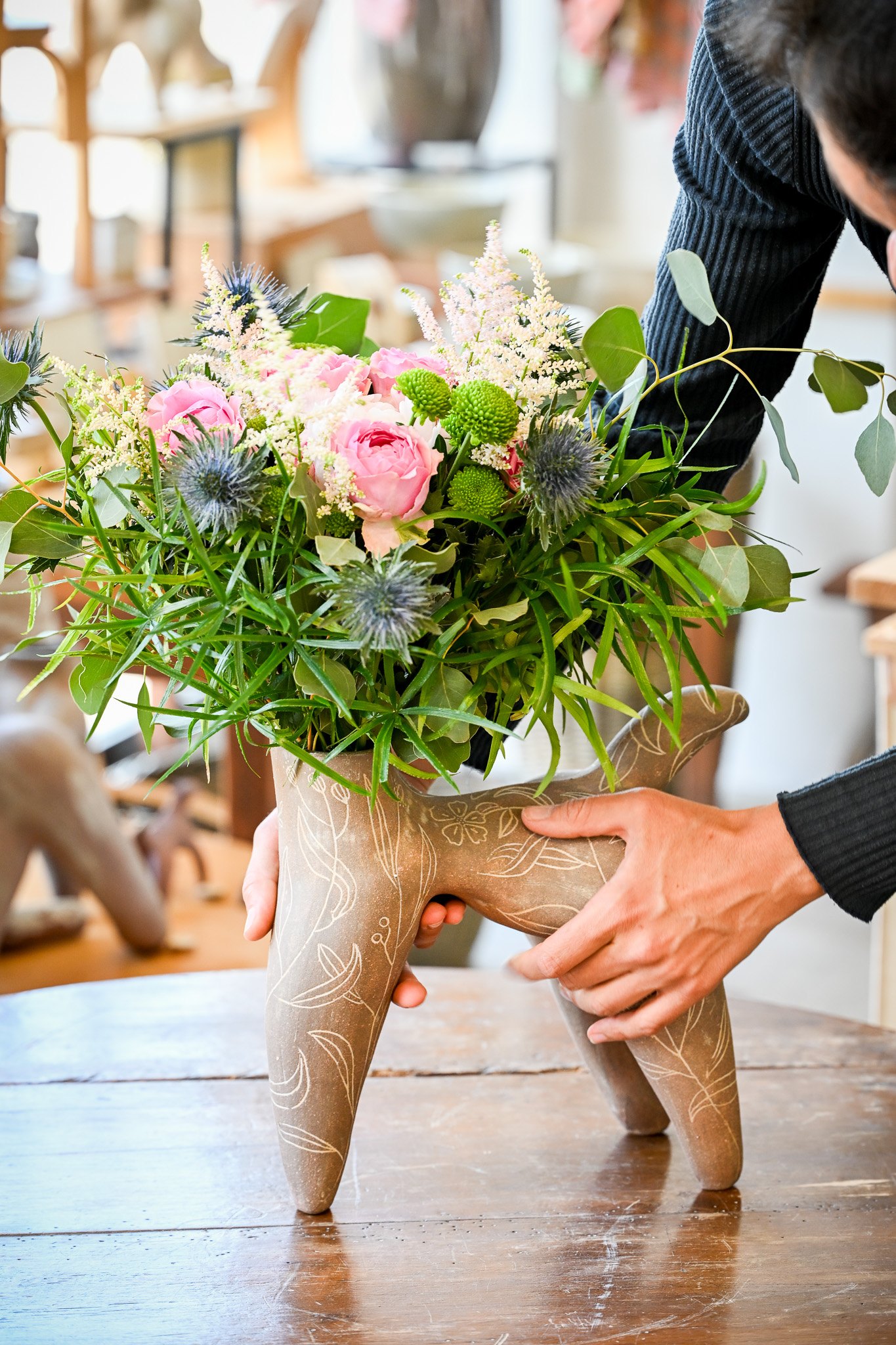 Deux mains posent la pièce aux feuillages sur une table en bois ; un bouquet de roses, de chardons bleus et d'eucalyptus sort de son col.