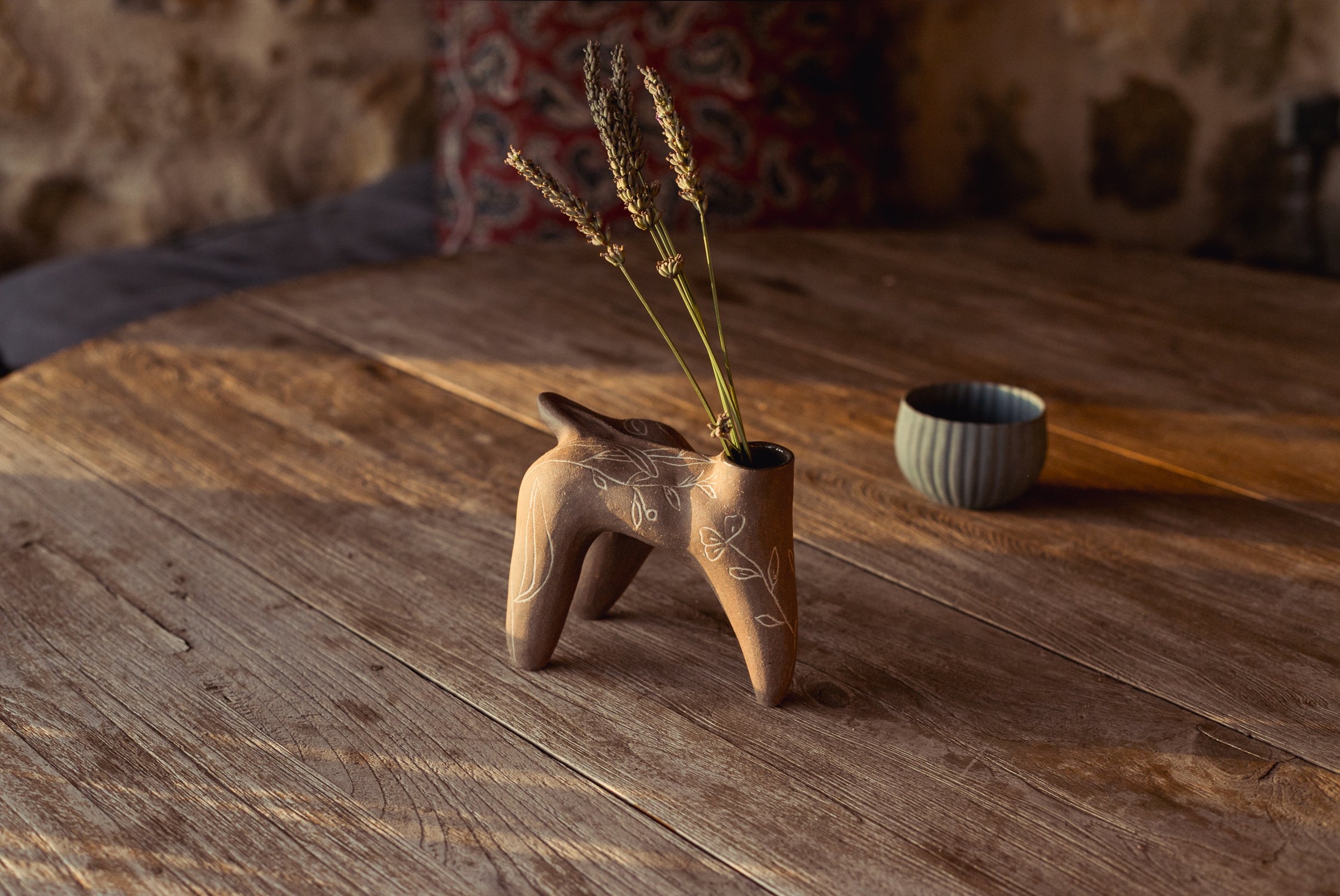 Pièce brune à quatre pieds portant des brins de lavande séchée, un bol clair à côté, sur une table en bois devant un mur de pierre, dans la lumière basse du soir.