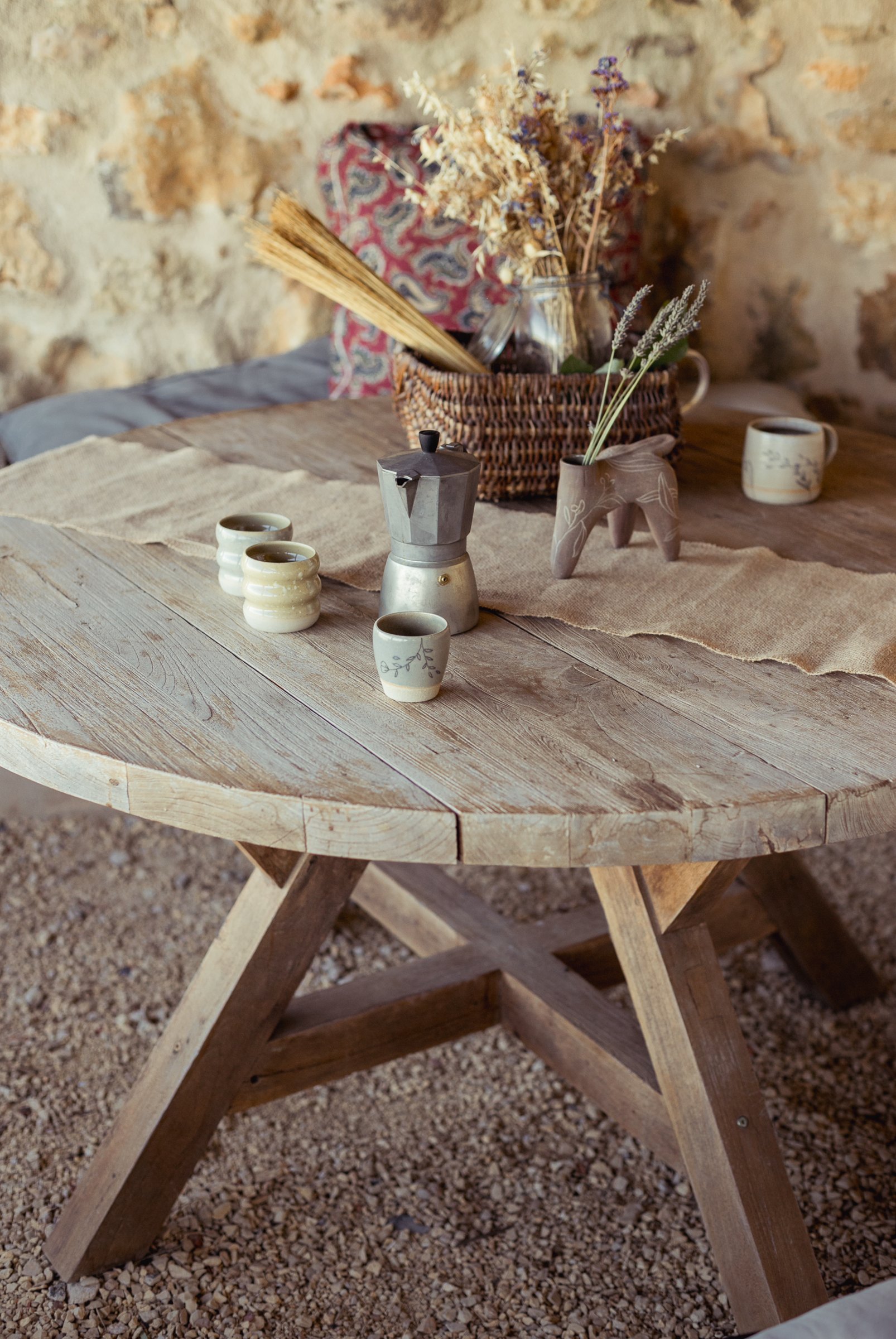 Table ronde en bois avec des tasses, un panier de fleurs séchées, devant un mur de pierre.